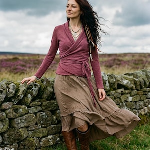 May include: A woman wearing a burgundy wrap top, a long brown skirt, and brown boots stands near a stone wall. Her long, dark hair is styled with braids. The outfit is suitable for outdoor wear.