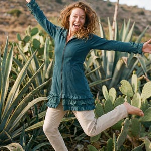 Puede incluir: Una chaqueta con capucha de color verde azulado con cremallera y dobladillo con volantes se usa con pantalones color beige. La persona está saltando frente a un fondo de plantas verdes y una montaña. La chaqueta tiene mangas largas con puños con volantes.
