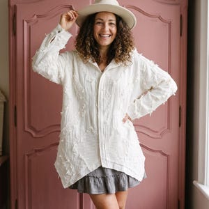 Könnte beinhalten: Eine weiße Langarmjacke mit strukturiertem Rautenmuster und ausgefransten Kanten, kombiniert mit einem grauen Minirock mit Rüschen und einem beigen Hut mit breiter Krempe. Die Jacke hat eine Knopfleiste vorne und eine lockere Passform.