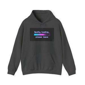 Puede incluir: Sudadera con capucha gris oscuro con bolsillo delantero y capucha. La parte delantera presenta un gráfico con el texto "Apathy loading... please leave" sobre una barra de carga azul y rosa.