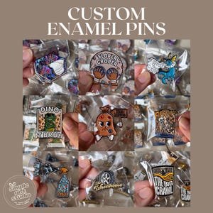 Puede incluir: Una colección de pines de esmalte personalizados en envases de plástico transparente. Los pines presentan varios diseños, incluyendo un dinosaurio, un esqueleto y texto que dice "SLOPER GROPER" y "THE BIG CRAWL". Los pines son coloridos y detallados.