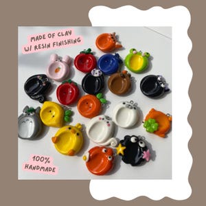 May include: A collection of colorful, handmade clay charms with resin finishing. The charms feature various designs, including animals, fruits, and other whimsical shapes. The text "MADE OF CLAY W/ RESIN FINISHING" and "100% HANDMADE" are visible.