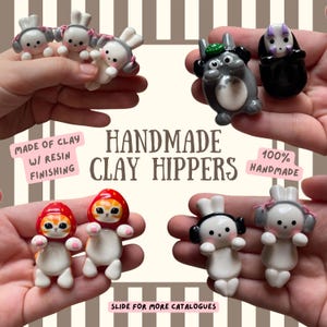Op de afbeelding: Een verzameling kleine, handgemaakte kleifiguren met harsafwerking, waaronder witte konijnen met roze strikjes, een grijs wezen met een groene hoed, een zwart figuur en oranje katten in aardbeikappen, met de tekst HANDMADE CLAY HIPPERS.