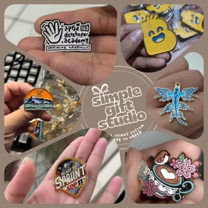 May include: A selection of vibrant enamel pins featuring diverse designs: a hand with text, a smiling yellow letter B, a mountain scene, a blue creature, a space-themed pin, and a teacup with flowers. These custom-made pins are on display.