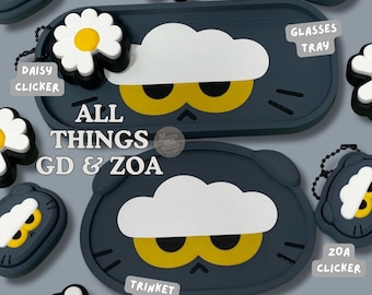 May include: A collection of dark gray silicone accessories featuring a white cloud and yellow eyes design. Items include a rectangular glasses tray, an oval trinket dish, and several small clickers with white daisy attachments. Text reads ALL THINGS GD & ZOA.