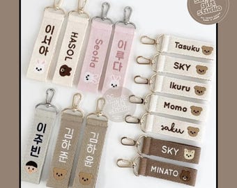 May include: A collection of personalized keychains in various colors, including beige, pink, and brown. Each keychain features a name or word in Korean or English, along with a small embroidered animal design. The keychains have silver-colored clasps and are labeled "Warmth Edition."