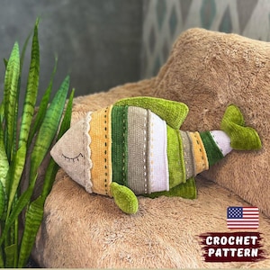 Könnte beinhalten: Ein handgehäkeltes Fischkissen in einem farbenfrohen Design. Der Fisch hat einen beigen Kopf mit geschlossenen Augen, einen gelb-grünen Körper und grüne Flossen. Der Text "CROCHET PATTERN" ist im Bild zu sehen.