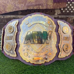 May include: A championship wrestling belt featuring a large central gold plate with the text AEW WORLD CHAMPION, surrounded by intricate filigree and embedded clear stones. The belt includes side plates with gold accents and a dark purple strap.
