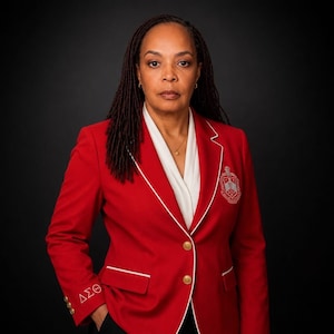 May include: A red blazer with white trim and gold buttons. The blazer features a crest on the chest and Greek letters on the sleeve. The person is wearing a white blouse underneath.