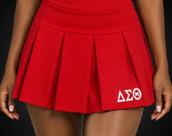 May include: A bright red pleated skirt featuring a high waistband and the white Greek letters Delta Sigma Theta printed on the lower right side. The skirt has a structured, flared silhouette with sharp vertical pleats throughout the fabric.