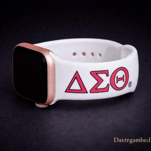 May include: A white Apple Watch band with a rose gold case. The band features the Greek letters Delta, Sigma, and Theta in red, with a matching red circle around the Theta. The watch is displayed on a dark surface.