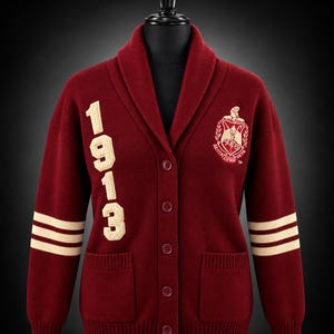 May include: A burgundy knit cardigan with a shawl collar and button front. The right side features the cream-colored text 1913, while the left side displays a cream-colored crest. Both sleeves have two cream stripes. Distinguished Legacy Co.