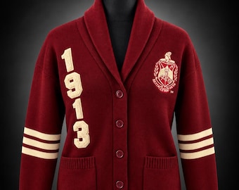 May include: A burgundy knit cardigan with a shawl collar and button front. The right side features the cream-colored text 1913, while the left side displays a cream-colored crest. Both sleeves have two cream stripes. Distinguished Legacy Co.