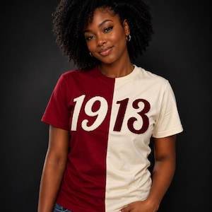 May include: A two-toned t-shirt with the year "1913" in white, featuring a burgundy and cream color block design. The model is wearing blue jeans. The shirt has short sleeves and a crew neck.