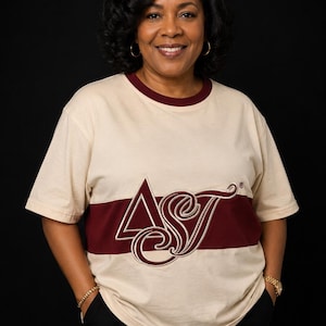 May include: A cream-colored short-sleeve t-shirt featuring a horizontal maroon stripe across the chest. The shirt displays the embroidered Greek letters Delta and Sigma, along with the letter Tau in a script font, centered over the maroon stripe.