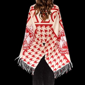 May include: Cream and red shawl with a houndstooth pattern and fringe. The shawl features Greek letters and crests in red. The letters "ΔΕΛΤΑ" and "ΘΕΤΑ" are visible. The shawl is draped over a person.