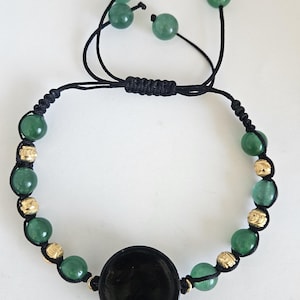 Puede incluir: Una pulsera de cordón negro con un dije central circular negro, alternando cuentas de piedra verde y pequeñas cuentas de metal dorado texturizado. La pulsera incluye un cierre deslizante ajustable con cuatro cuentas de piedra verde colgantes.