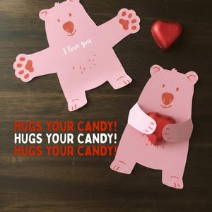 Könnte beinhalten: Rosa bärenförmige Valentinstag-Kartenhalter mit dem Text "I love you" und "HUGS YOUR CANDY!". Ein Bär hält ein rotes herzförmiges Bonbon. Der andere Bär hat ausgestreckte Arme. Ein rotes herzförmiges Bonbon ist ebenfalls vorhanden.