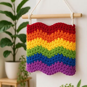 May include: A colourful, handmade crochet wall hanging in a rainbow design. The piece has a wavy edge and is made up of red, orange, yellow, green, blue, and purple bands. It hangs from a wooden dowel with white rope. Potted plants are in the background.