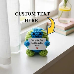 May include: A crocheted Earth-shaped plush toy in blue and green, holding a sign that reads "You Make The World A Better Place" with a red heart. The text "CUSTOM TEXT HERE" is above the toy. The toy is on a white surface.
