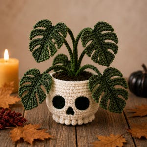 Op de afbeelding: Een gehaakte monstera-plant met donkergroene bladeren in een crèmekleurige schedelvormige pot. De schedel heeft diepe zwarte oogkassen en een driehoekige neusholte, met een getextureerd oppervlak dat de botstructuur nabootst.