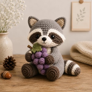 May include: A crocheted raccoon plush toy featuring gray and white yarn, black eye patches, and a striped tail. The raccoon is holding a cluster of purple crocheted grapes with a single green leaf. The toy has dark brown paws and ears.