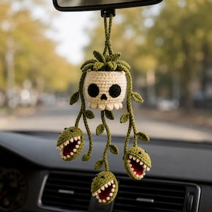 May include: A crocheted car hanging ornament featuring a cream-colored skull-shaped planter with green leafy vines trailing downwards. Three small green carnivorous plant heads with open mouths, white teeth, and red interiors hang from the vines.