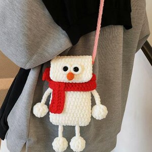 May include: A white, crochet snowman-shaped crossbody bag with black button eyes, an orange nose, and a red scarf. The bag has a pink strap and pom-pom feet and hands. The bag is worn over a gray sweatshirt.