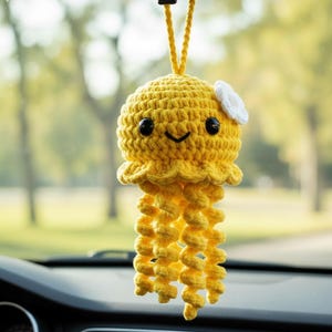 May include: A yellow crocheted jellyfish car charm with a smiling face, black eyes, and a small white flower. The jellyfish has long, spiraling tentacles and is suspended by a yellow string, adding a whimsical touch to any vehicle's interior.