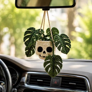 May include: A crocheted hanging ornament featuring a cream-colored skull-shaped planter containing dark green monstera leaves. The plant is suspended by a cream-colored cord, designed to hang from a vehicle rearview mirror, showcasing detailed yarn textures and intricate leaf patterns.