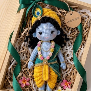 May include: A crochet doll with light blue skin, black hair, and a yellow turban featuring a peacock feather. The doll wears a yellow dhoti, a green shawl, and a floral garland. A round tag with the text Handmade is attached to the box.