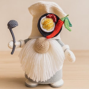 May include: A handmade crochet gnome chef wearing a white hat and apron. The gnome has a long white beard and is holding a ladle. A plate of ramen and a red chilli pepper are attached to the gnome's chest.