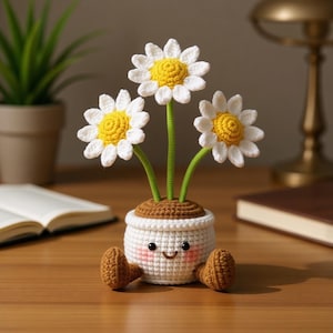 Könnte beinhalten: Ein gehäkeltes Gänseblümchen-Blumenarrangement in einem weißen Topf mit einem lächelnden Gesicht und braunen Füßen. Die Gänseblümchen haben weiße Blütenblätter und gelbe Zentren mit grünen Stielen. Der Topf steht auf einer Holzoberfläche.