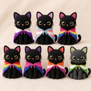 May include: Seven crocheted cat figurines with large black eyes and whiskers, each wearing a small cape featuring different coloured stripes. The cats are primarily black, with one grey cat, and the capes display various pride flag colour patterns.