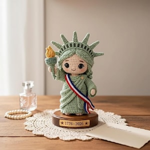 May include: A crocheted Statue of Liberty doll in shades of green, holding a golden torch. The doll has a red, white, and blue sash and is mounted on a wooden base with the text "1776-2026".