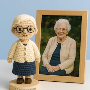 May include: A crocheted doll of an elderly woman wearing glasses, a cream-colored cardigan, and a blue skirt, standing next to a framed photograph of a smiling elderly woman. The doll is on a wooden base with the text "In loving memory".