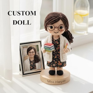 Può includere: Bambola personalizzata all'uncinetto di una donna con occhiali, un vestito floreale e un cardigan bianco, che tiene libri e una matita. La bambola è su una base di legno con il nome "Miss Harper". È presente anche una foto incorniciata della donna. Il testo "CUSTOM DOLL" è sopra.