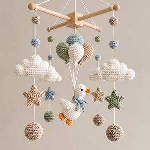 May include: A hanging nursery mobile featuring a central white crocheted duck with an orange beak and feet, suspended by strings attached to three crocheted balloons in sage green, white, and beige. Surrounding elements include white clouds, stars, and spheres in muted blue, green, and beige tones, all attached to a light wooden cross frame.