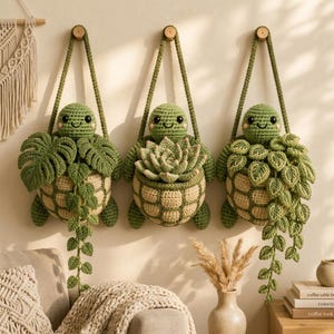 Puede incluir: Tres adornos de pared de tortugas tejidas a ganchillo en tonos verde y beige. Cada tortuga tiene un caparazón que contiene una planta diferente tejida: una monstera, una suculenta y una enredadera. Tienen ojos negros y mejillas rosas.
