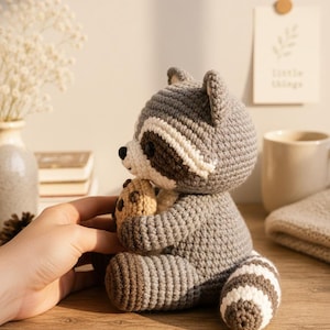 May include: A crocheted raccoon toy made of grey, white, and dark brown yarn. The raccoon has a striped tail, a black eye mask, and is holding a small, round, tan-colored cookie. The toy is crafted with visible stitches and has a soft, plush appearance.