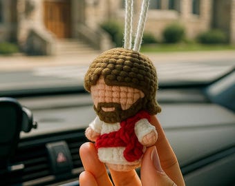 May include: A crocheted Jesus car charm. The charm features a brown-haired figure with a beard, wearing a white robe and a red sash. The charm is suspended by white cords, and is held in front of a church.