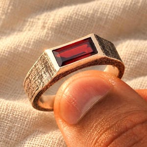 May include: A silver-toned ring featuring a rectangular, deep red gemstone set horizontally. The metal band has a dark, textured, and weathered finish, contrasting with the smooth, polished surface of the vibrant red stone centered on the top.