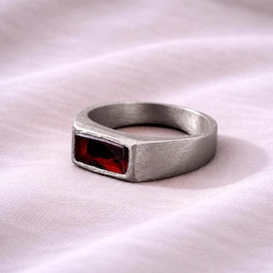 Puede incluir: Un anillo de tono plateado con acabado cepillado y mate que presenta una única piedra preciosa rectangular de color rojo intenso engastada horizontalmente en el centro. La banda tiene un perfil ancho y plano con bordes rectos y uniformes.