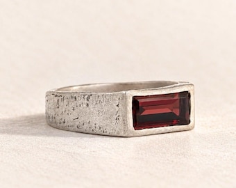 May include: A silver-toned ring featuring a rectangular, deep red gemstone set horizontally. The metal band has a textured, weathered finish, providing a matte appearance that contrasts with the smooth, polished surface of the dark red stone.