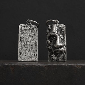 May include: Two rectangular silver-toned metal pendants with a weathered finish. One pendant features a sculpted face with a beard and mustache, while the other displays a textured surface with the engraved text AMOR FATI at the bottom.