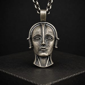 Pode incluir: Um pingente de metal prateado em forma de cabeça humana estilizada com traços faciais definidos, incluindo olhos amendoados, nariz reto e lábios finos. O pingente tem um acabamento texturizado e desgastado, pendurado em uma corrente grossa de tom prateado.