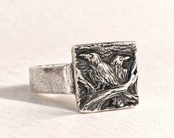 May include: A silver-toned ring featuring a square face with a relief design of two birds perched on a branch. The metal has a textured, oxidized finish, giving it a weathered appearance. The band is wide and also displays a brushed, rustic texture.
