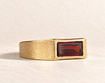 May include: A gold ring with a brushed matte finish featuring a single rectangular baguette-cut deep red garnet gemstone set horizontally in the centre of the band. The metal has a textured surface and a warm yellow tone throughout.