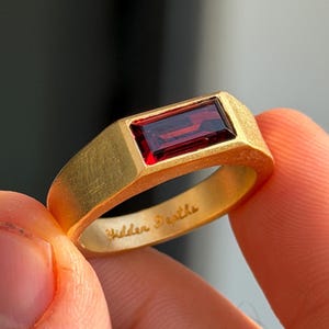 Könnte beinhalten: Ein goldfarbener Ring mit mattem, gebürstetem Finish und einem horizontal gefassten, rechteckigen, dunkelroten Edelstein. Auf der Innenseite des Rings ist der kursive Schriftzug Hidden Depths eingraviert, der auf der Metalloberfläche gut sichtbar ist.