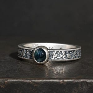 May include: A silver ring featuring a central round, dark blue-green stone set in a bezel. The band is crafted with an oxidized finish and displays a continuous relief pattern of small, stylized figures and shapes encircling the entire circumference.